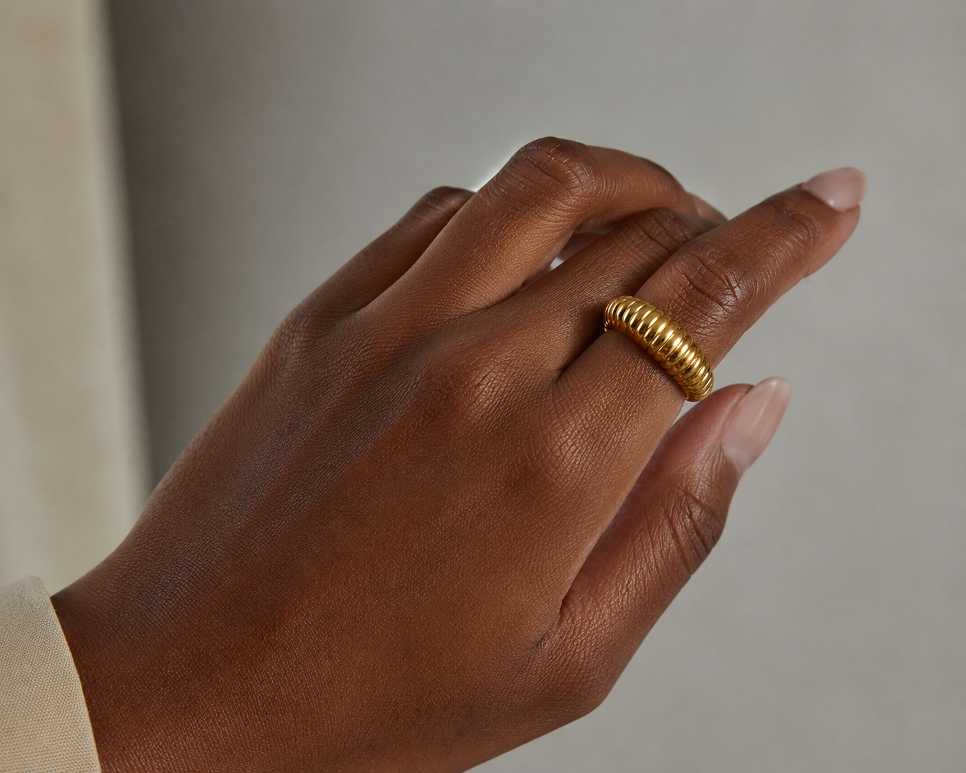 A close-up of a hand wearing a textured gold ring on one of the fingers, set against a soft neutral gray background.