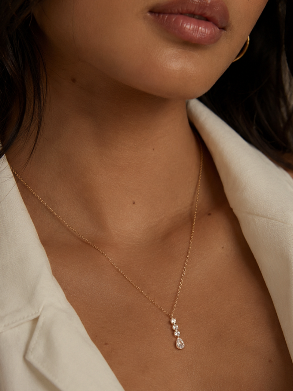 A close-up lifestyle shot of a model wearing a delicate gold chain necklace with a small teardrop pendant set with clear stones, styled with an open white shirt.