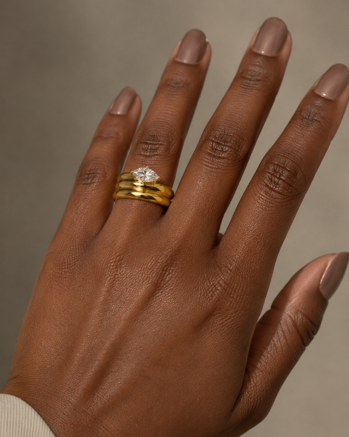 A close-up of a hand wearing three stacked rings, including a yellow gold band with a small clear gemstone, against a soft neutral background.
