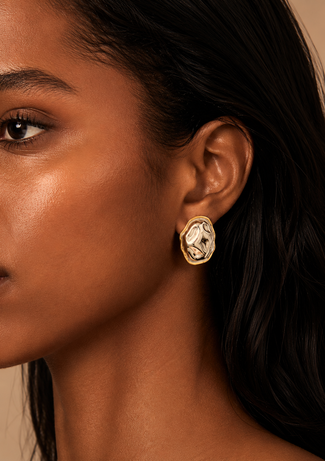 A close-up side profile of a model wearing a gold oval stud earring with a textured, abstract in silver and gold .