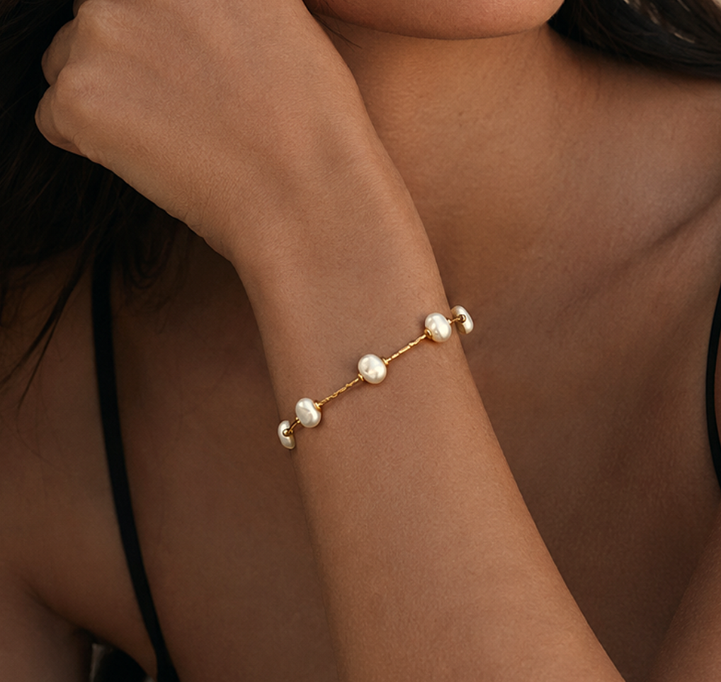 A close-up of a woman wearing a delicate gold chain bracelet with small modern white pearls, styled with a black spaghetti-strap top.